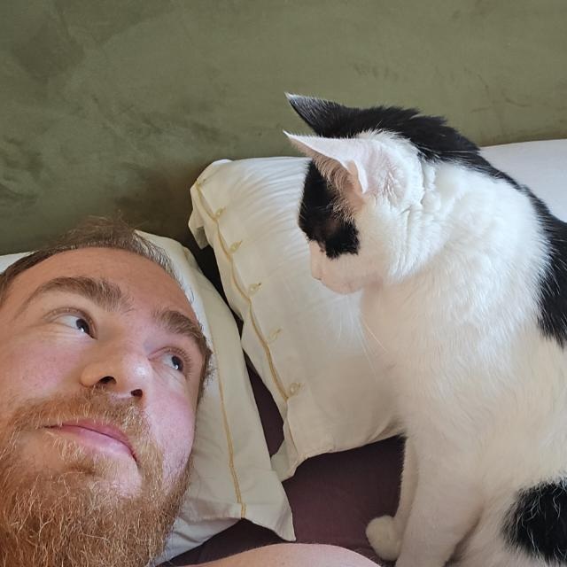Carlo Ruggiero lying down, looking up at a black and white cat sitting beside him